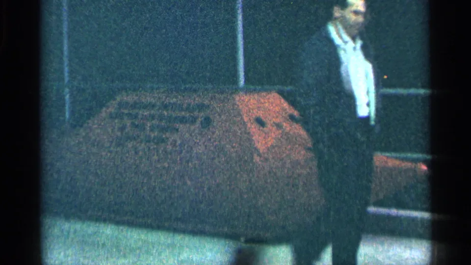 1964 Niagara Falls frame showing an adult man in a dark suit beside a rust-colored barrel at a display area.