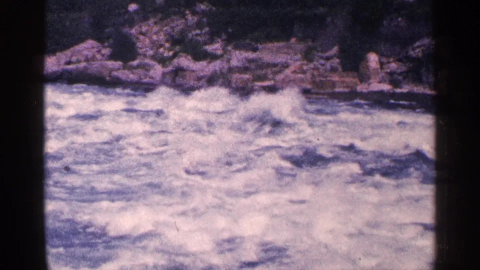 A visitor-shot 8mm view shows turbulent Niagara Falls area rapids moving across the frame below a rocky shoreline.