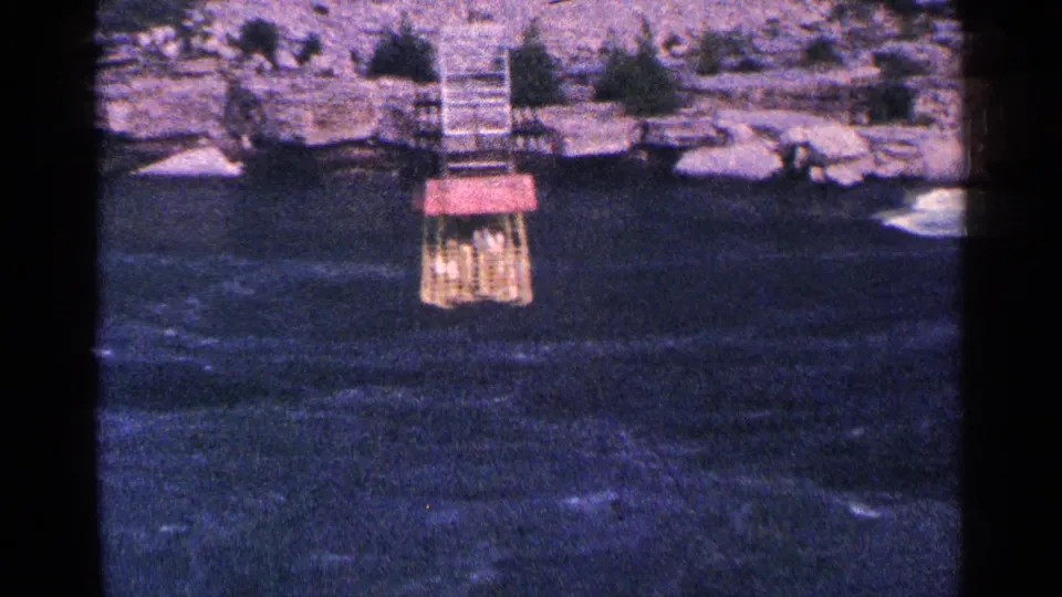 Distant 1964 Niagara Falls river view with a small red-roofed structure on the water, steps, rocks, and scattered visitors ashore.
