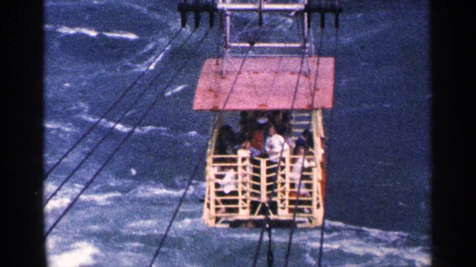 1964 Niagara Falls area home-movie frame showing an open-sided cable car with passengers crossing above rushing blue water.