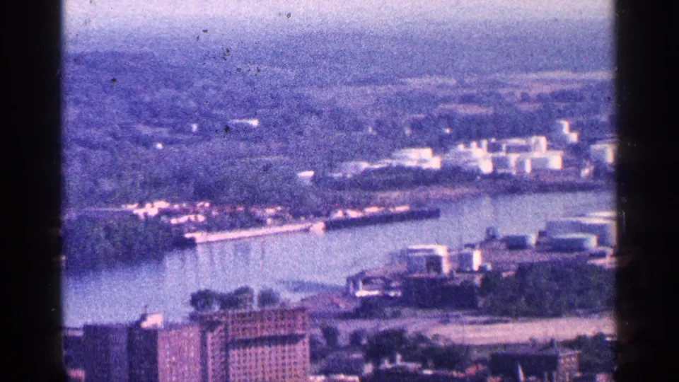 Elevated 1964 home-movie view across water near Niagara Falls, with buildings, docks or shoreline structures, and hazy distance.
