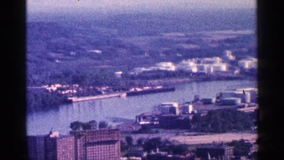 1964 8mm view likely near Niagara Falls, Canada, showing a riverfront industrial area, city blocks, and distant tree-lined terrain.