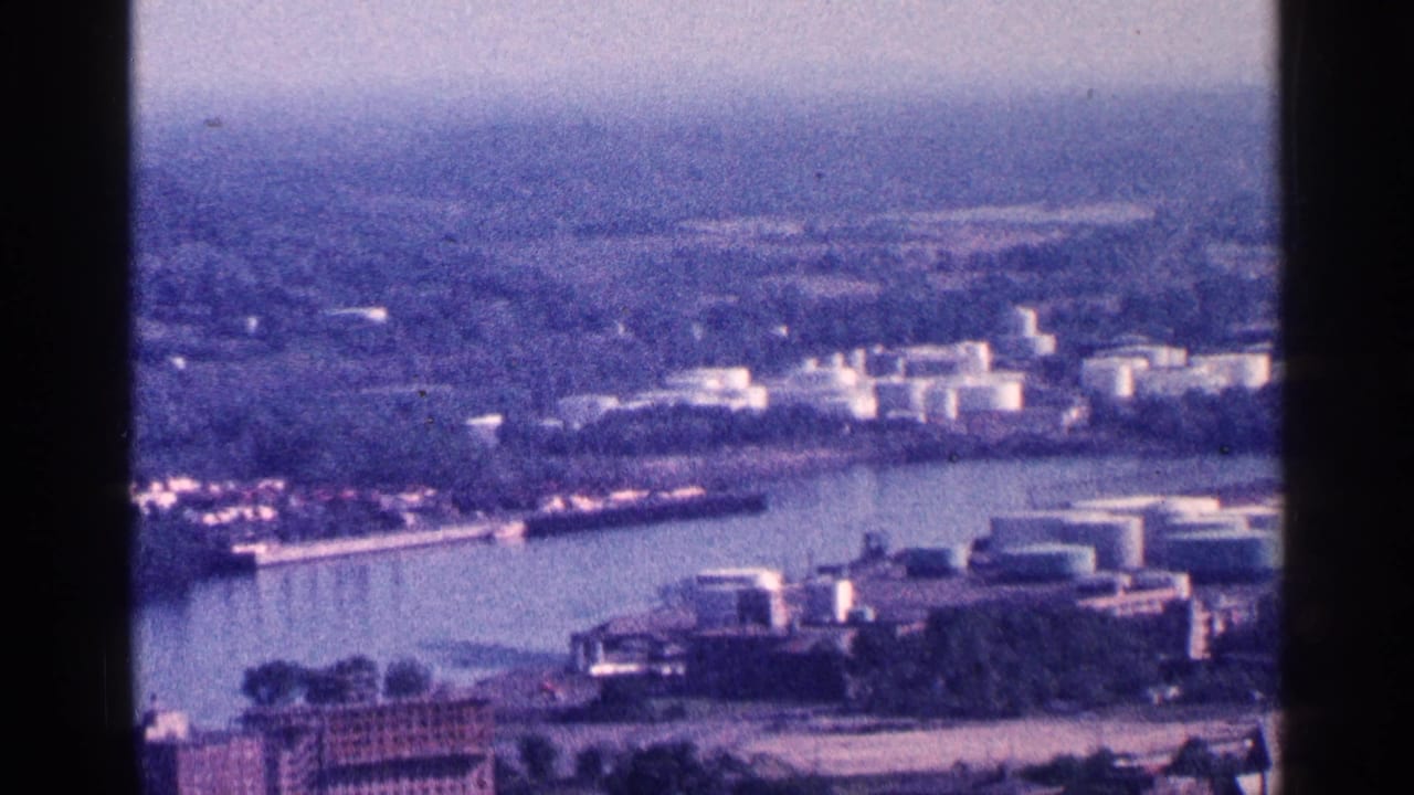 Likely Niagara Falls area riverfront in 1964, with industrial structures, storage tanks, and a hazy city landscape beyond the water.