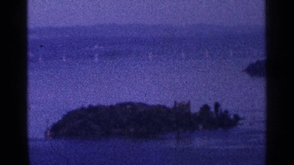Blue-tinted 1964 8mm view of a small wooded island in the Hudson River area, with hazy distant shore behind it.