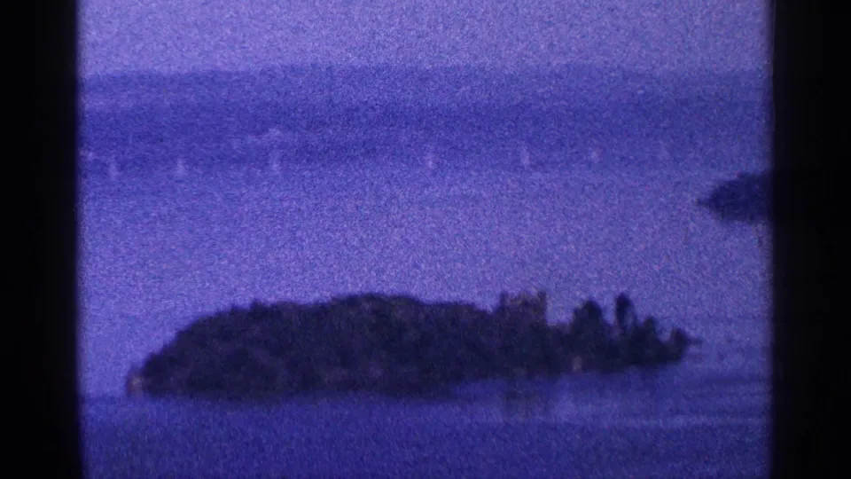 1964 8mm frame near Niagara Falls, Canada, showing a small wooded island across purple-shifted water with a soft far shoreline.
