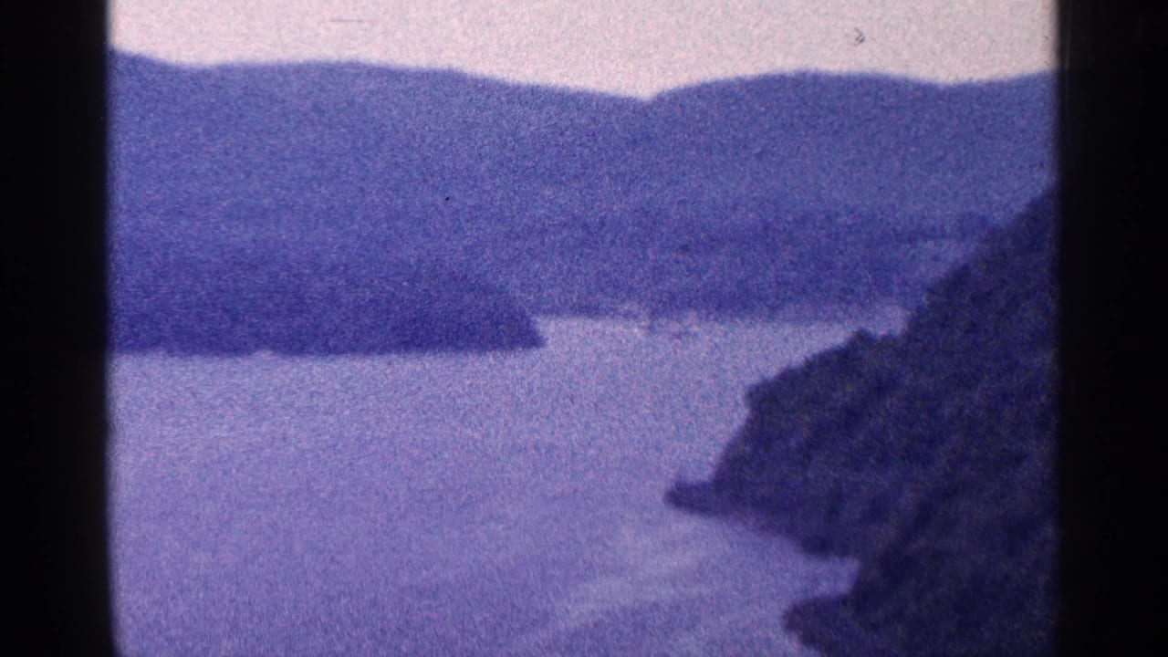 1964 8mm home-movie frame showing calm water, dark wooded headlands, distant hills, and a hazy sky near the supplied Niagara Falls, Canada location.