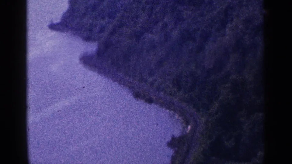 1964 8mm frame looking along a narrow stretch of water beside a forested hillside, with soft focus and purple color cast.