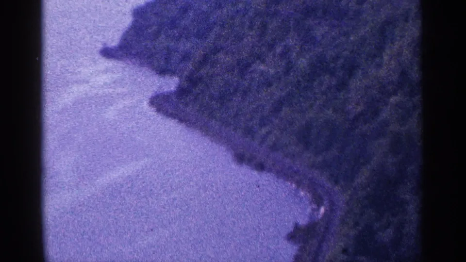 1964 8mm home-movie frame with a closer view of rippled water, a wooded point, and soft shoreline detail in grainy purple tones.