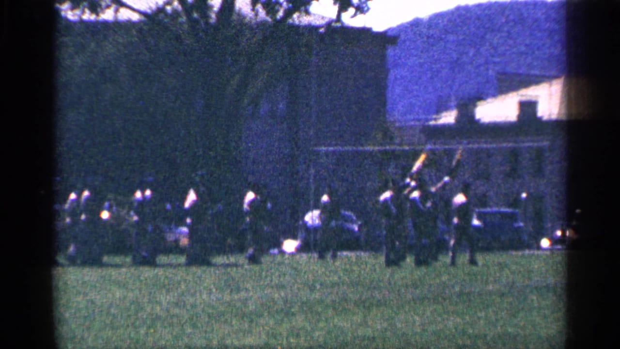 1964 Niagara Falls-area home-movie frame showing distant uniformed adults moving in formation across grass near trees and a large building.