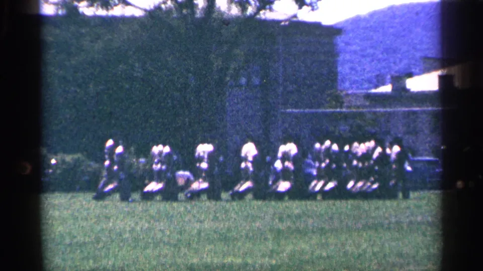 1964 Niagara Falls-area home-movie frame with uniformed adults clustered on a field near a dark vehicle or equipment shape.