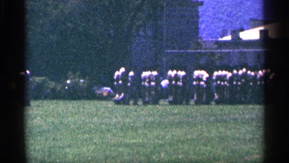Distant 1964 Canada frame showing a line of uniformed adults on grass, partly obscured by soft focus and dark film borders.