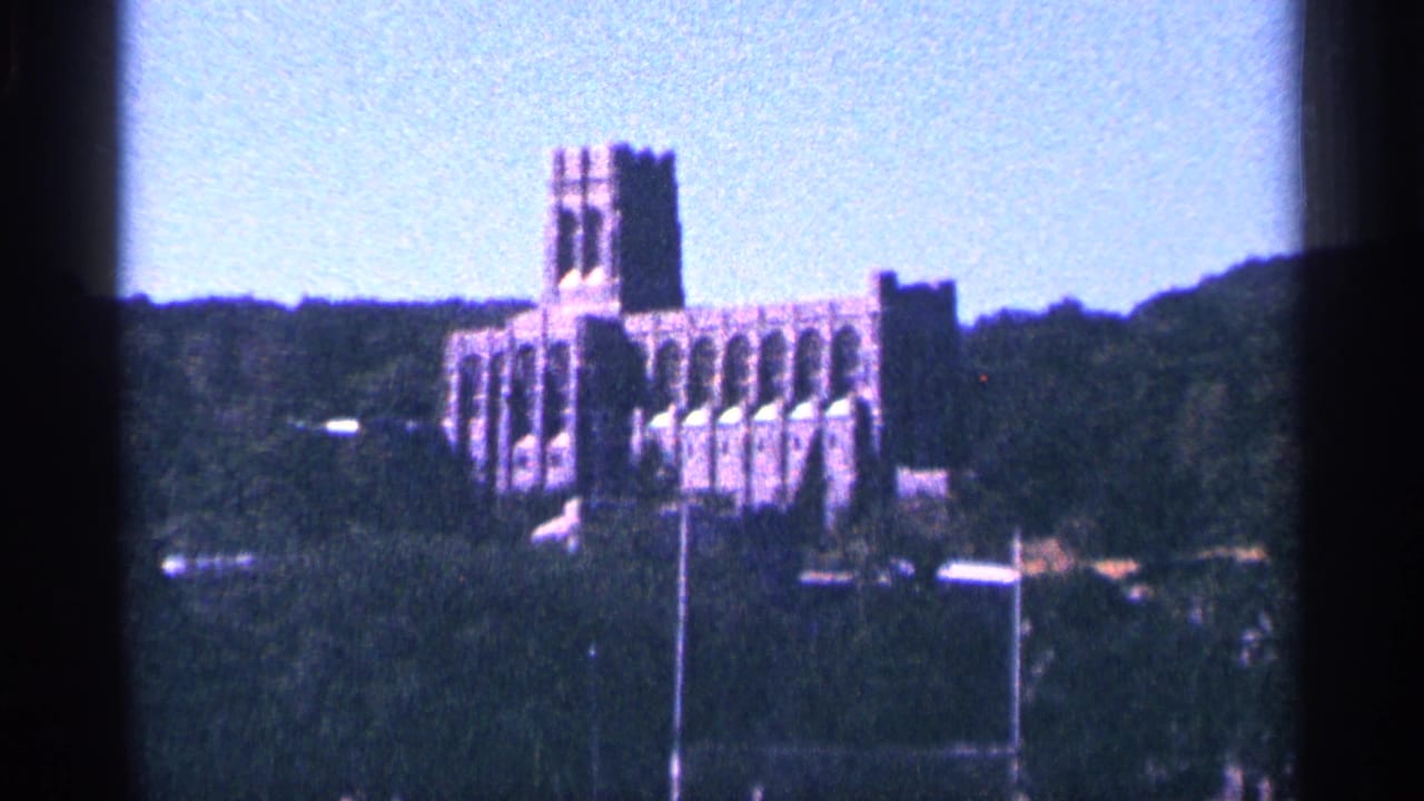 Distant gothic-style brick building on a wooded hill above a field.