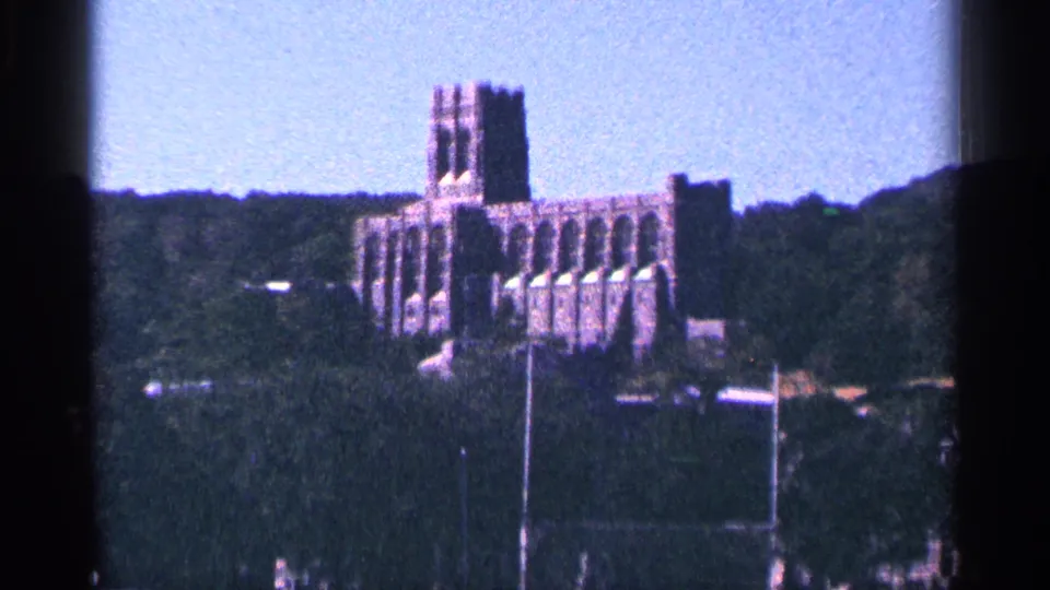 1964 8mm frame of the distant brick building on a wooded rise, with a field and goal-like structures in the lower foreground.
