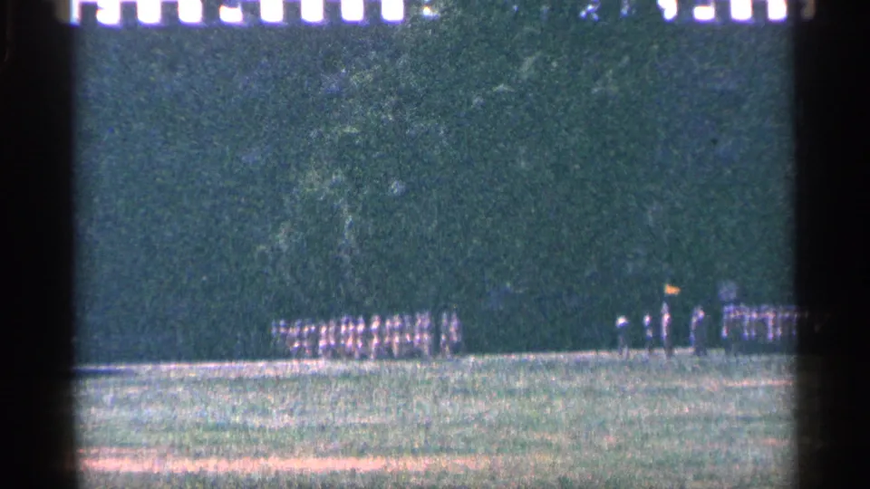 1964 Niagara Falls Canada home-movie frame showing a small group of distant people in formation on a field below a wooded area.