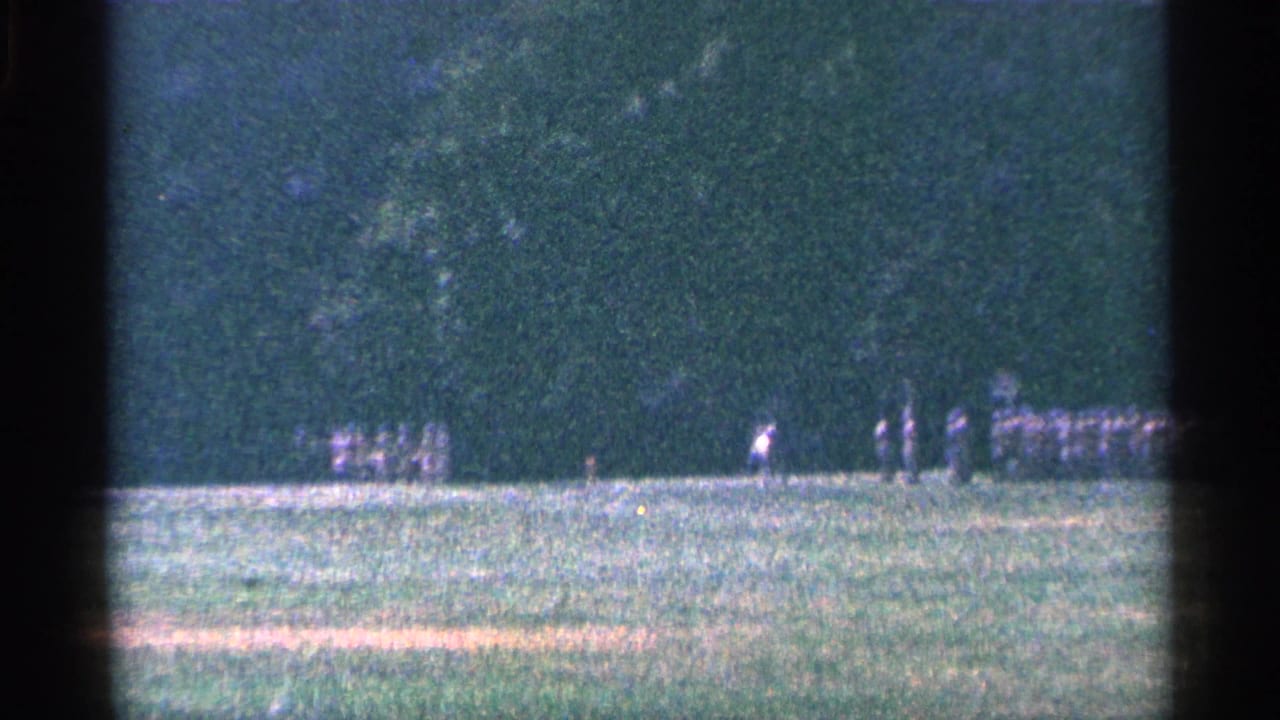 Small groups of uniformed men move across a grassy field with trees in the background.
