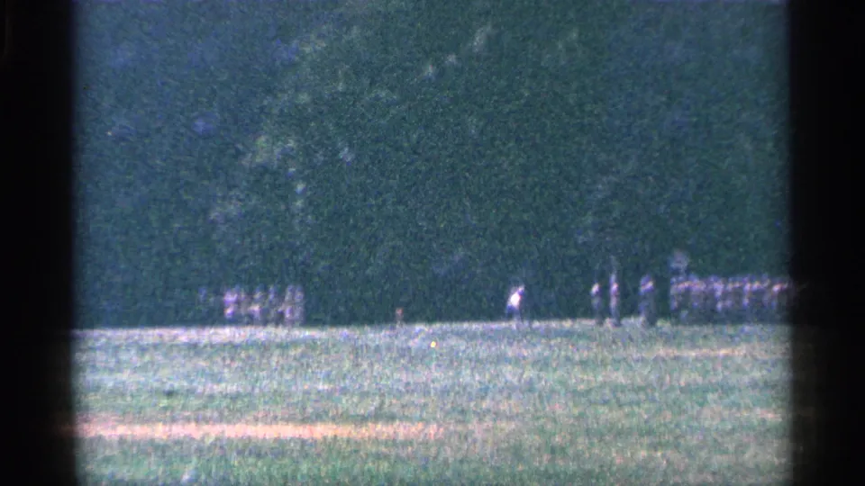 Distant uniformed figures continue moving across a grassy field in 1964 Niagara Falls, Canada home-movie footage.