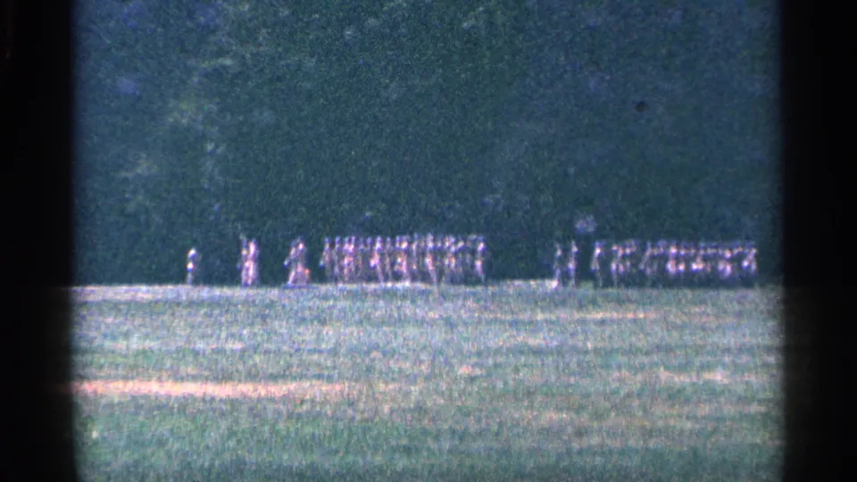 1964 home-movie frame of distant uniformed men in separate formations on a grassy field near a tree line.