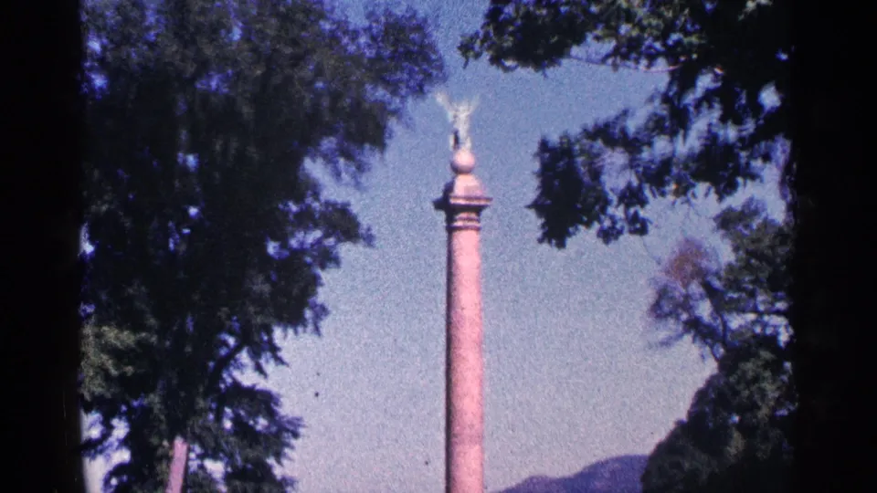 Closer 8mm frame of the winged statue atop a column, with trees and low hills visible in the Niagara Falls, Canada clip metadata.