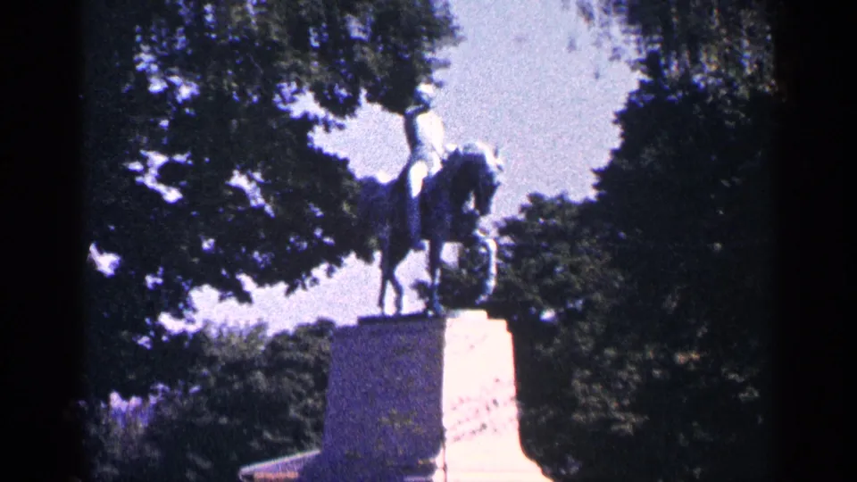 1964 8mm home-movie frame showing an equestrian statue on a stone pedestal in a tree-lined public setting.