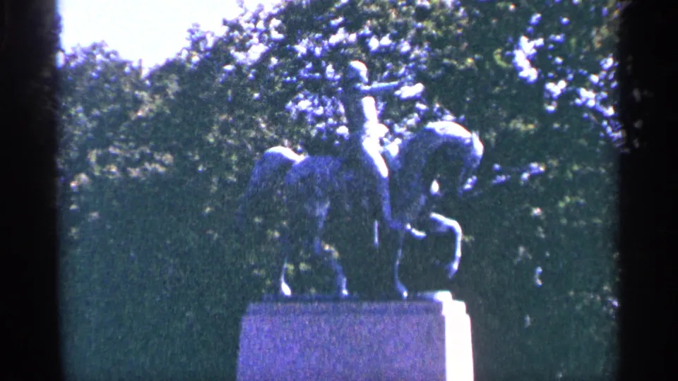 The 8mm footage holds on a public equestrian statue, with leafy trees and blue-tinted grainy exposure around the monument.