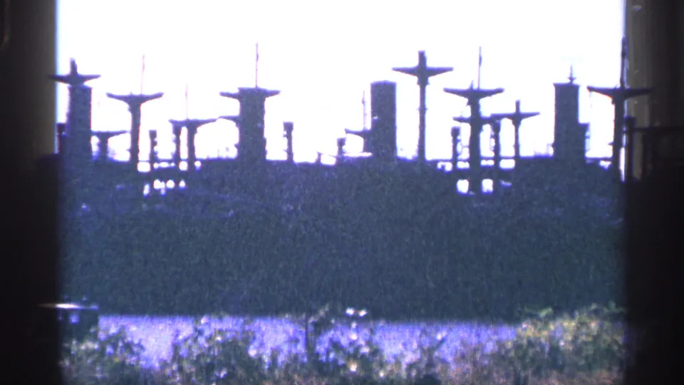 1964 8mm frame showing silhouetted ship masts, funnels, shoreline brush, and water near the reserve fleet setting.
