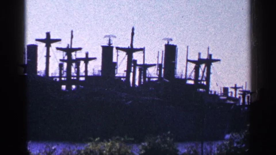 1964 home-movie frame showing the ship's dark profile with cross-shaped mast structures and a blue dusk cast.