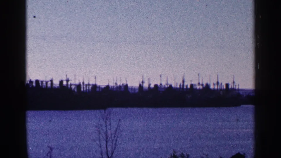 Blue-tinted 8mm frame of a distant Niagara Falls, Canada waterfront, with masts or dock structures silhouetted beyond the water.