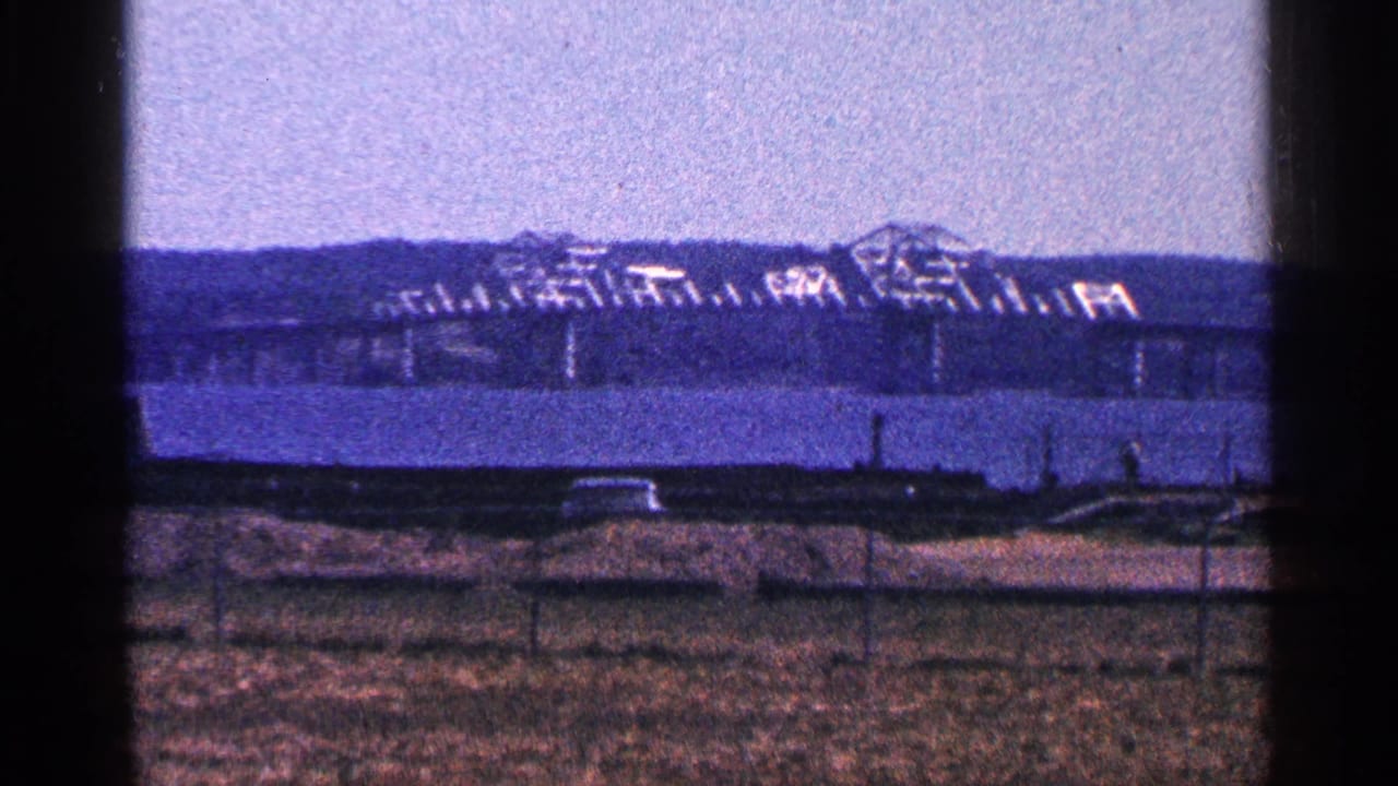1964 Niagara Falls, Canada home-movie frame showing a distant bridge and industrial structure across a broad river from a grassy fenced foreground.