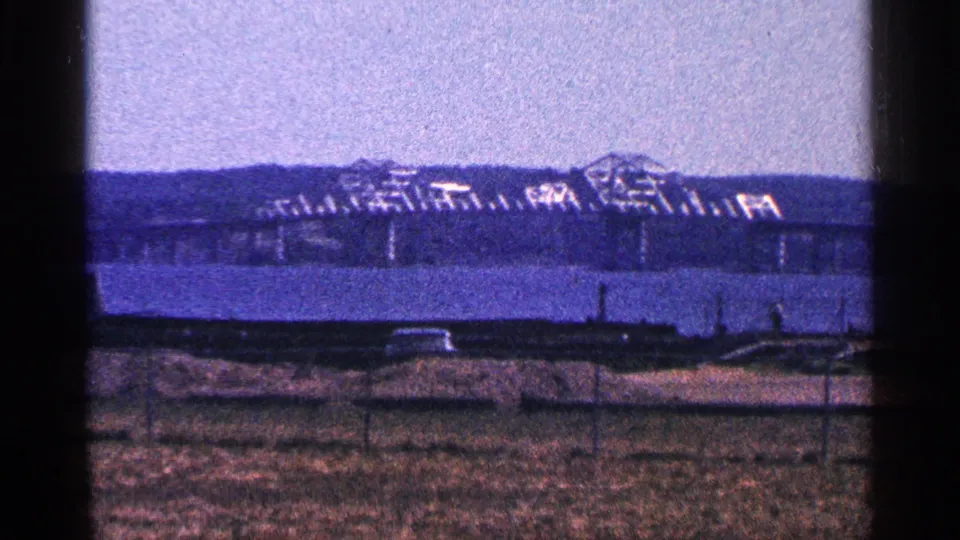 1964 Niagara Falls, Canada home-movie frame showing a distant bridge and industrial structure across a broad river from a grassy fenced foreground.