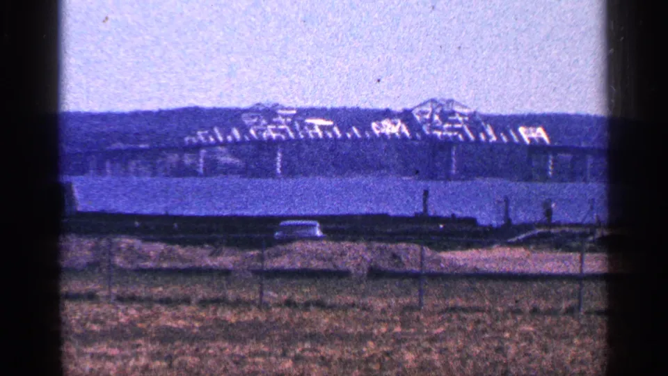 Distant 1964 Niagara Falls, Canada river view with a bridge and industrial structure beyond a grassy fenced area and roadside edge.