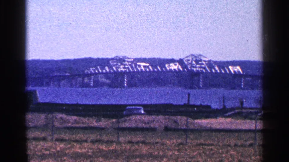 Soft-focus 1964 Niagara Falls, Canada frame showing grass, fence, river, bridge, and a distant industrial structure on the opposite bank.
