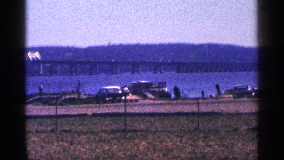 1964 Niagara Falls, Canada home-movie frame with parked cars and small people by the river, facing a distant bridge and hillside.