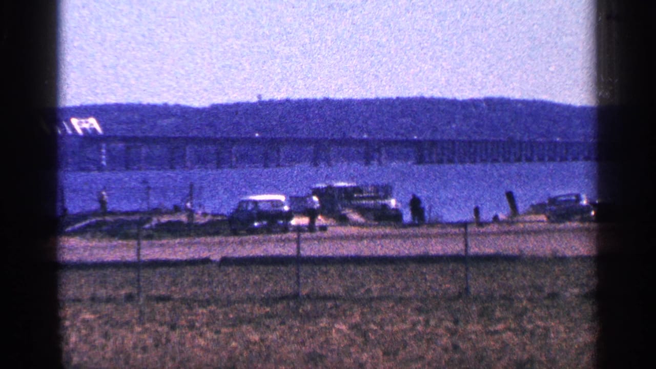 1964 8mm travel footage from Canada showing a waterfront road, long bridge, parked classic cars, fence, and hazy hills.