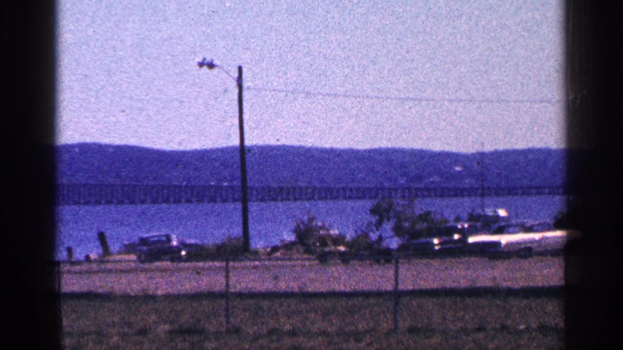 Waterfront road with parked cars, a utility pole, fence, bridge, and distant hills.