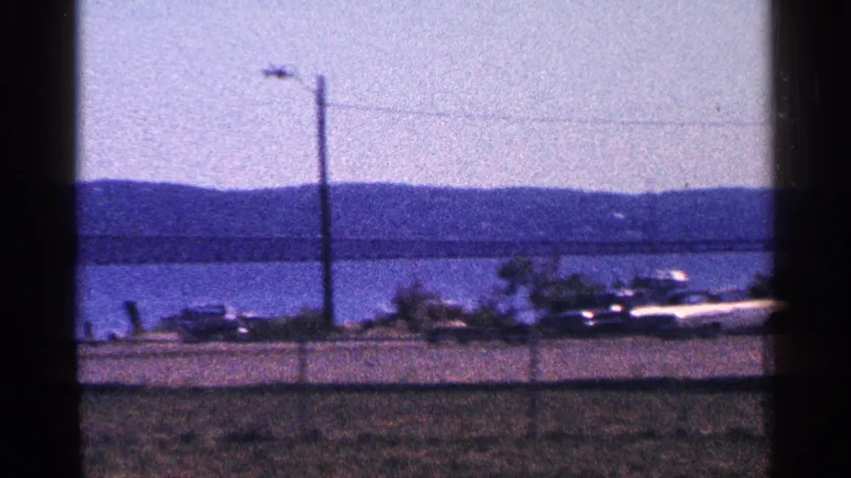 1964 8mm view of classic cars parked near a waterside road, with fence, utility pole, bridge, and hazy hills in the background.
