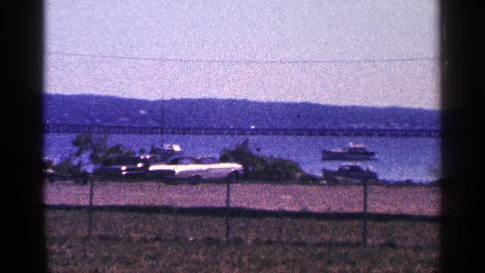 1964 Canada waterfront frame showing parked cars, chain-link fence, long bridge, distant hills, and a small boat on the water.