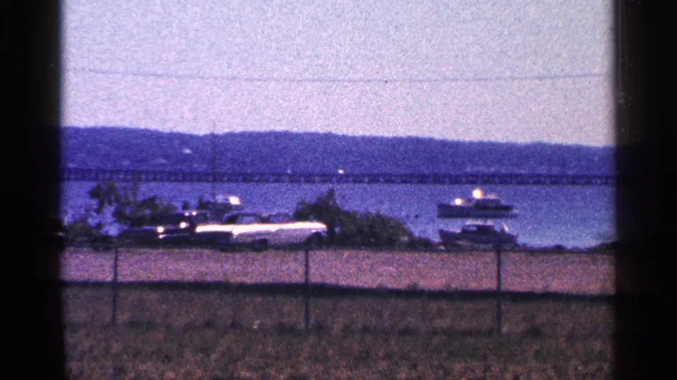 1964 8mm view near Niagara Falls, Canada, showing boats by shore, parked cars, fencing, water, hills, and a long bridge.
