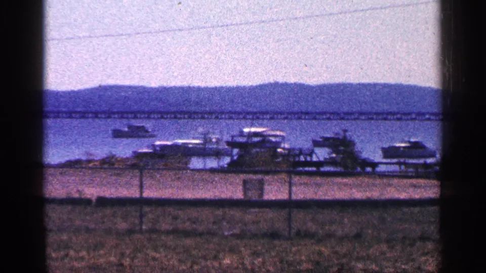1964 8mm shoreline view with docked boats, open water, distant hills, and a long bridge near Niagara Falls, Canada.