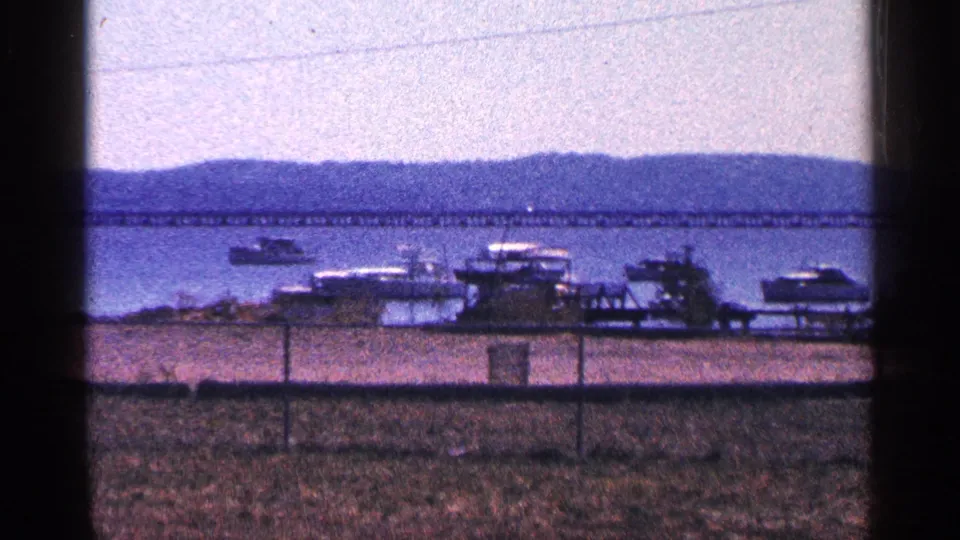 1964 home-movie frame of boats at a waterfront pier, fence in front, and a long bridge over water near Niagara Falls, Canada.