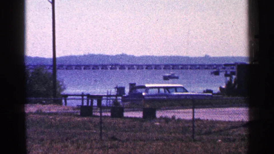 1964 8mm view near Niagara Falls, Canada, with a parked station wagon, waterfront fence, boats, water, hills, and bridge.