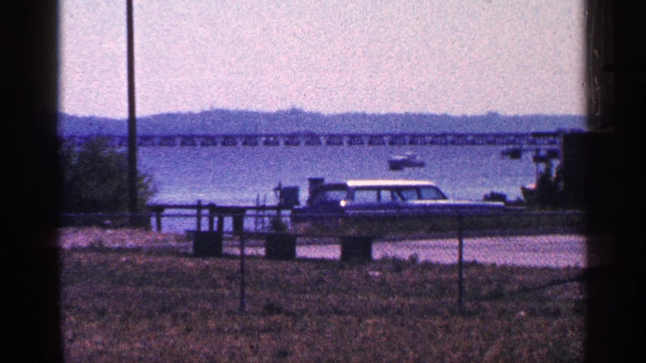 1964 8mm travel frame showing a station wagon beside a waterfront, with a long bridge, hazy hills, and a small boat beyond.