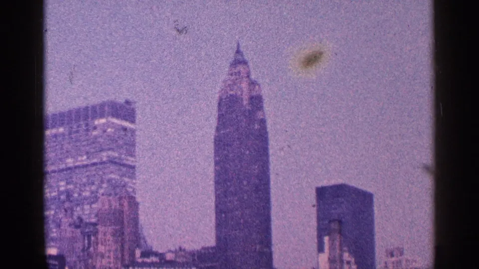 1964 8mm frame of a city skyline with a prominent spire-topped tower, older masonry building, and modern high-rise blocks.