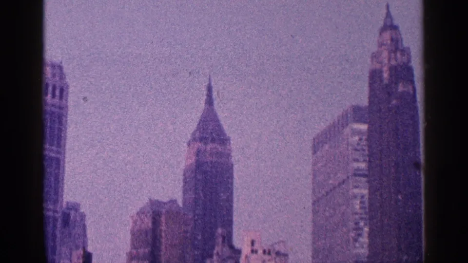 1964 8mm skyline frame showing tall downtown buildings, including a pointed-roof tower and adjacent high-rise structures.