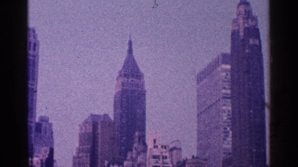 1964 8mm frame of a dense urban skyline with multiple towers, including spire-topped buildings and block-like high-rises.