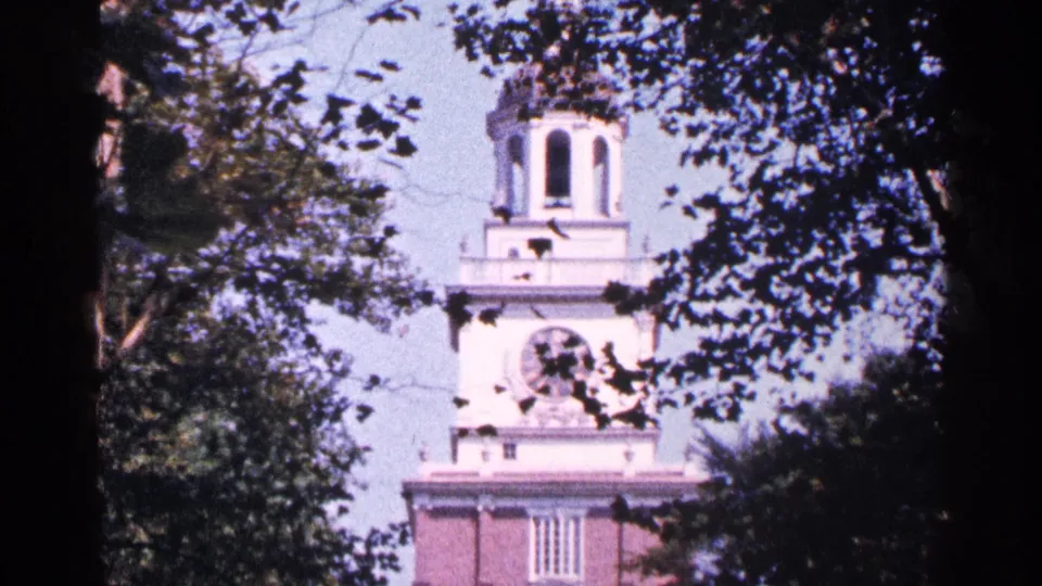 1964 Pennsylvania 8mm frame of a red brick historic-looking building with a white clock tower seen through leafy trees.