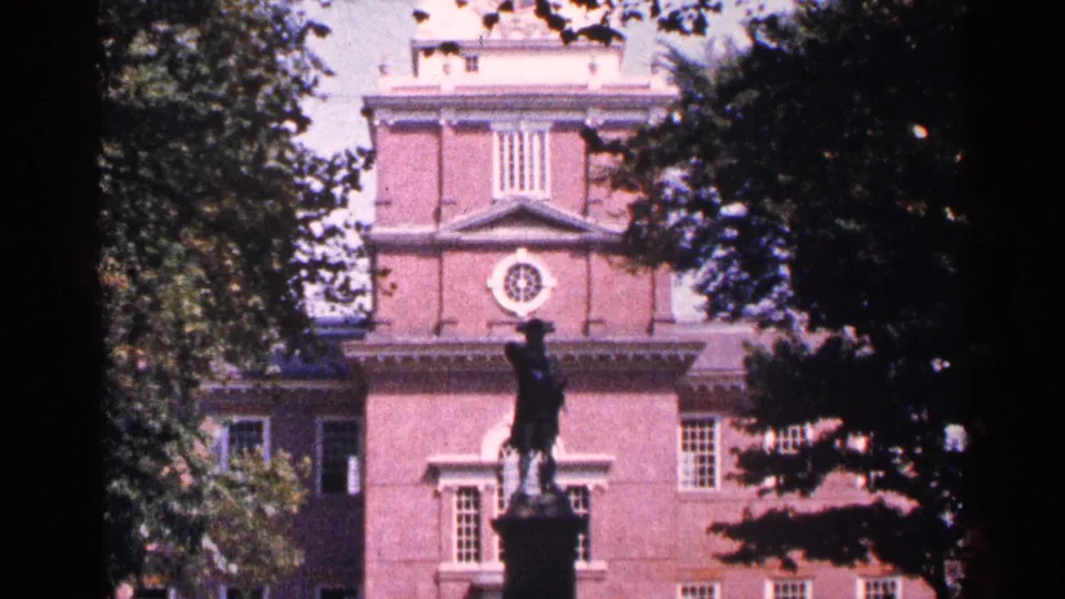 1964 Pennsylvania 8mm frame showing a statue centered before a red brick historic-looking building with white trim.