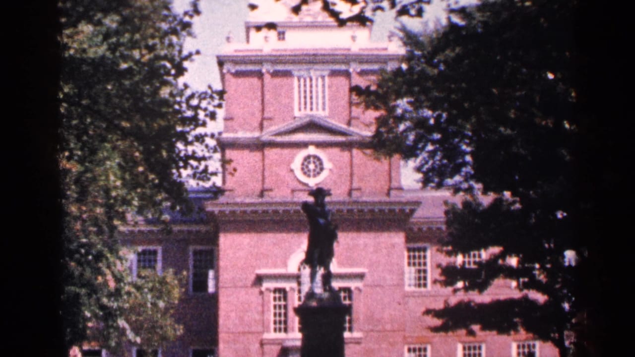 1964 home-movie view of Independence Hall in Philadelphia, partly obscured by trees, with a statue silhouetted in front.