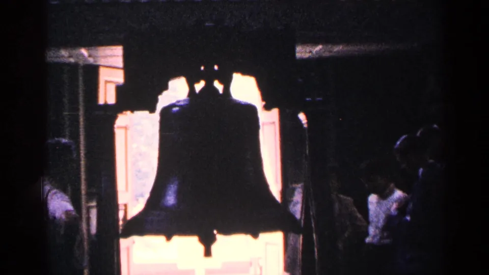 1964 Pennsylvania home-movie frame showing the Liberty Bell silhouetted against overexposed light, with partially visible visitors nearby.