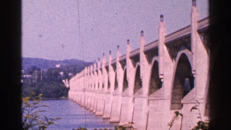 1964 Pennsylvania travel frame showing a long pale arched bridge over a river, with wooded hills and blue sky beyond.