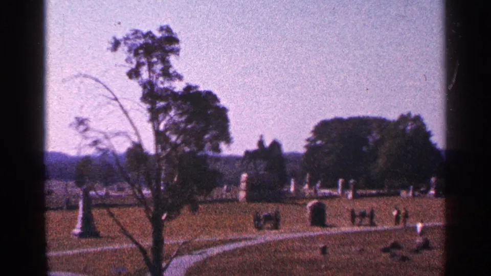 1964 Pennsylvania 8mm view of Gettysburg cemetery grounds with trees, paths, scattered headstones, and a distant monument.